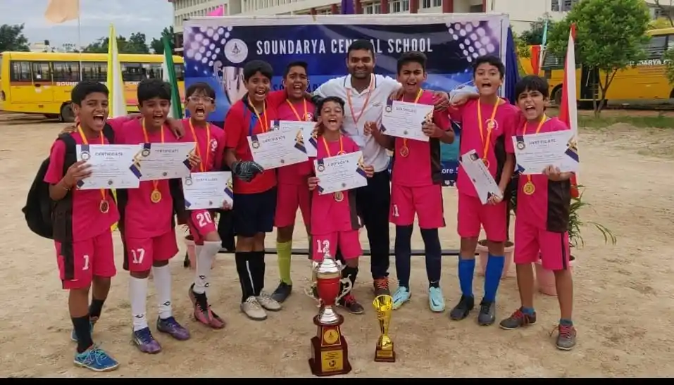 A group of young athletes in pink uniforms holding certificates and medals, posing with trophies in an outdoor setting.